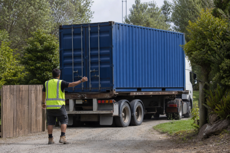 Blue shipping container arriving on a flatbed truck at a job site with a worker guiding delivery